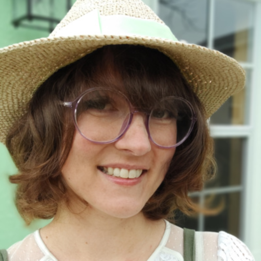 Photo of SednaWoo, a smiling person with short curly brown hair wearing a woven sun hat and round purple glasses.