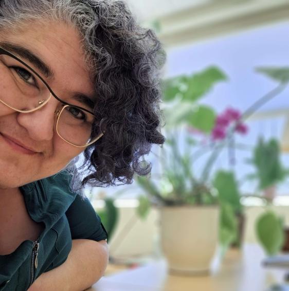 A smiling woman leans into the image frame on the left edge, smiling at the camera. She has dark brunet hair, curly, and touched with purple and natural grey. In the background, slightly out of focus, as houseplants. The whole space is brightly lit by sunlight.