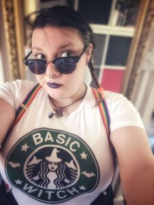 Selfie of a woman in sunglasses and rainbow suspenders. Her shirt imitates the Starbucks logo and says "Basic Witch."