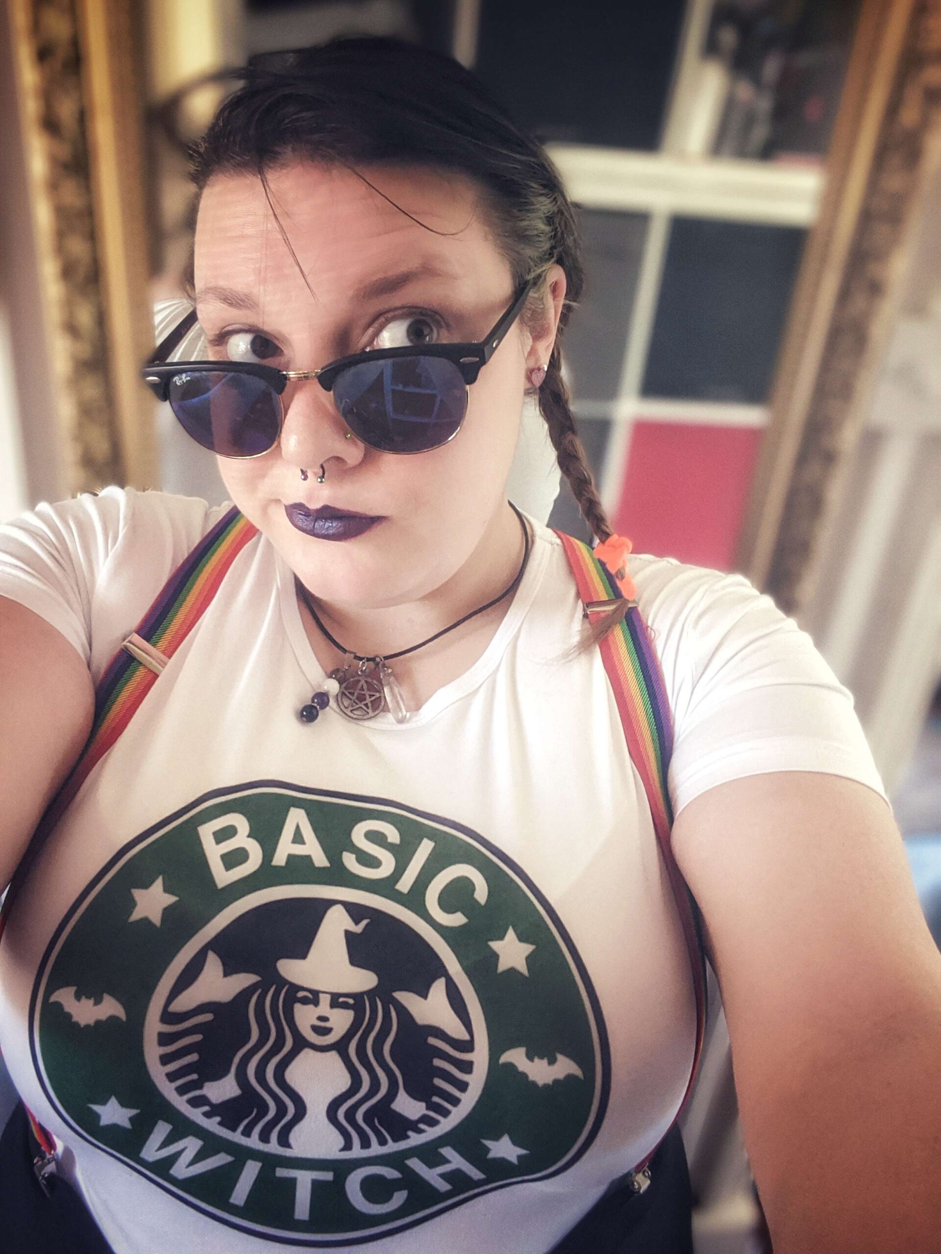 Selfie of a woman in sunglasses and rainbow suspenders. Her shirt imitates the Starbucks logo and says "Basic Witch."