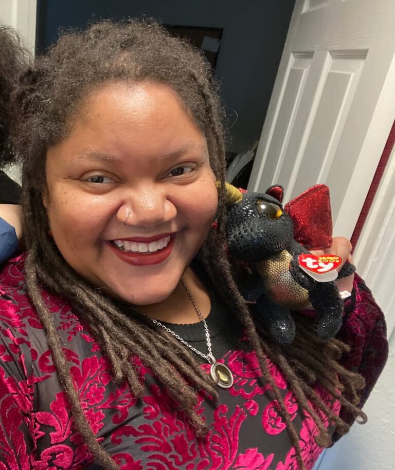 A selfie of a smiling woman with dreadlocks in a red and black velvet top. She's holding a black dragon TY beanie baby stuffed toy.