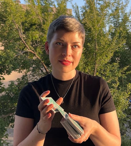 Person with short hair wearing a black shirt, red lipstick, shuffling tarot cards, standing outside in the golden sun.