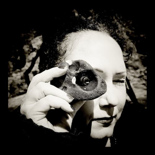 A black and white photo of a person with dark hair, lipstick, and painted nails holding up a hagstone and peering at the camera through it.