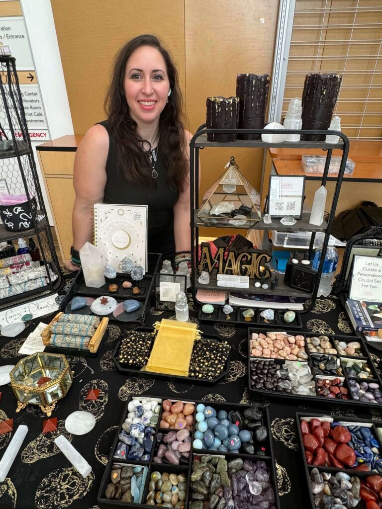 An assortment of crystals on a black shop table. A woman with long dark hair smiles behind the table.