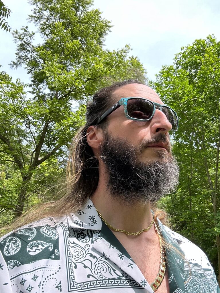 A man standing outside in sunglasses. He has long hair and a curly beard and gold chains around his neck.