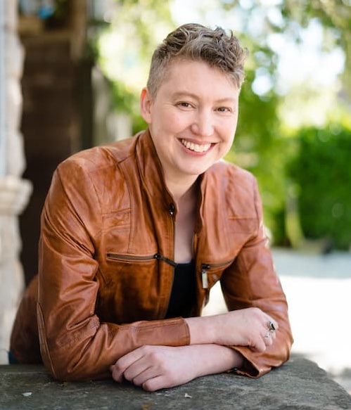 Photo of Lane, a light-skinned person with very short graying dark blonde hair that is shaved on the sides and wavy on top, smiling and wearing a brown leather jacket over a black tank top. They are leaning forward with arms crossed on a flat stone surface with a blurred urban environment with green trees in the background.