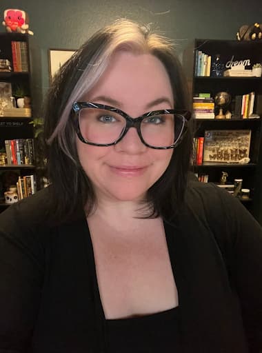 A person with shoulder-length dark hair with one light streak on the front, big black glasses, and dark clothing, standing in front of bookshelves.