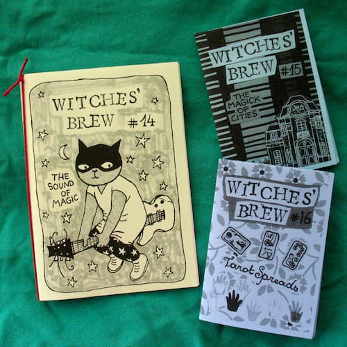 Three zines spread on a dark green tablecloth. All three are titled “Witches’ Brew”. The biggest zine features a drawing of a cat riding a guitar like a broomstick next to the text “the sound of magic” and the issue number “14”. One of the smaller zines, issue number 15, is subtitled “the magic of cities” and has an image of an old building. The other smaller zine, number 16, is subtitled “tarot spreads”, with images of hands and tarot cards.