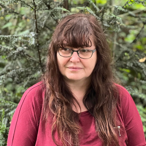 A person with long brown hair, glasses, and a maroon long sleeve shirt, standing in front of an evergreen bush and blurred green in the background.