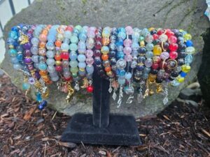 24 bracelets hang from a cylindrical black velvet stand made. They are outside on dark brown woody mulch and in front of a small grey lichen-spotted rock. The lighting is bright but not in direct sunlight. Bracelets are made from glass beads with metallic silver and gold charms including animals, plants and geometric shapes. Bracelets left to right: blue and gold, blue and silver, purple and gold, grey and silver, orange and blue, blue and grey, orange and yellow, red and grey, blue and green, blue and pink, pink blue and silver, red and black, orange and gold, black and silver, blue and silver, pink and silver, grey and silver, green and gold, brown and green, red black and yellow, dark red and crystalline bright red, red and gold, black and silver, blue and yellow.