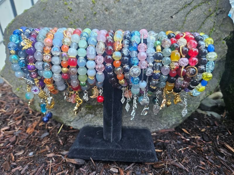 24 bracelets hang from a cylindrical black velvet stand made. They are outside on dark brown woody mulch and in front of a small grey lichen-spotted rock. The lighting is bright but not in direct sunlight. Bracelets are made from glass beads with metallic silver and gold charms including animals, plants and geometric shapes. Bracelets left to right: blue and gold, blue and silver, purple and gold, grey and silver, orange and blue, blue and grey, orange and yellow, red and grey, blue and green, blue and pink, pink blue and silver, red and black, orange and gold, black and silver, blue and silver, pink and silver, grey and silver, green and gold, brown and green, red black and yellow, dark red and crystalline bright red, red and gold, black and silver, blue and yellow.