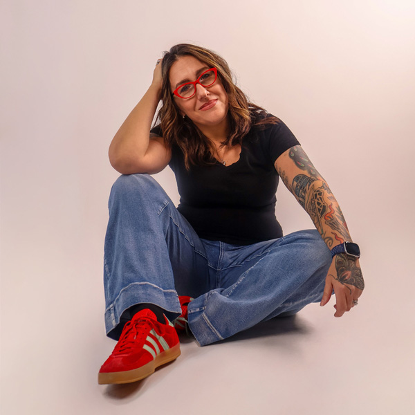 Adrienne sits cross-legged in front of a light background, wearing red glasses, a black shirt, blue jeans, and red Adidas sneakers. She has light skin, shoulder-length brown hair, and a tattooed sleeve on one arm.